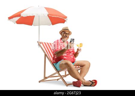 Reifer Mann mit einem Cocktail und einem Mobiltelefon auf einem Strandstuhl unter einem Sonnenschirm auf weißem Hintergrund isoliert sitzen Stockfoto