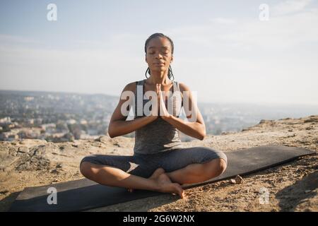 Lächelnde amerikanerin, die in Lotusposition sitzt, die Augen geschlossen und mit namastigen Gesten mit Stadtbild auf dem Hintergrund. Schwarze schlanke Dame, die Yoga auf einem hohen Hügel praktiziert. Stockfoto