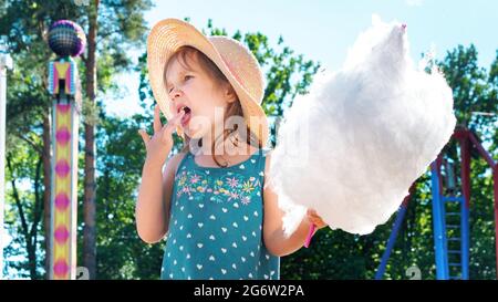 Nettes kleines Mädchen, das an einem Sommertag ein riesiges Stück Zuckerwatte in einem Vergnügungspark isst. Happy Childhood Konzept. Vorschulmädchen isst Zuckerwatte wi Stockfoto