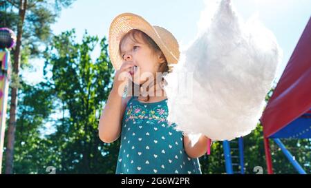 Das Kind leckt klebrige Finger. Ein niedliches kleines Mädchen isst ein riesiges Stück Zuckerwatte. Spazieren Sie mit Kindern im Park im Urlaub an einem sonnigen Tag. Stockfoto