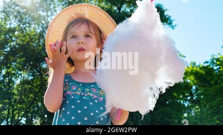 Das Mädchen schaut erstaunt und überrascht in den Vergnügungspark. Ein Kind isst an einem sonnigen Sommertag Zuckerwatte und leckt klebrige Finger. Gehen Sie mit ch Stockfoto