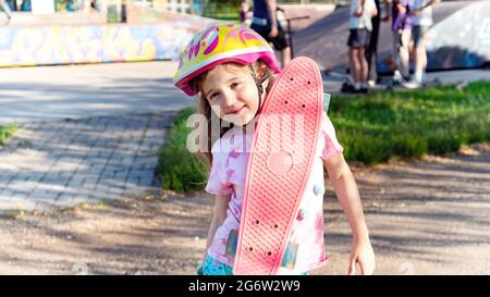 Ein süßes Mädchen, das vor dem Hintergrund eines Skateparks ein Penny Board auf der Schulter hält. Portrait eines sportlichen Mädchens in einem Helm an einem Sommertag am Stockfoto