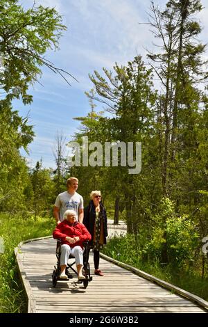 Großmutter, Tochter und ihr Sohn genießen einen Spaziergang in einem Naturgebiet mit Promenade, Enkel schieben Rollstuhl für die Mobilität, Baileys Harbor, WI, USA Stockfoto