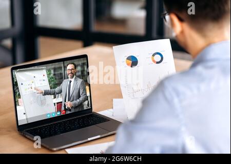 Über die Schulter schauen Sie, wie ein kaukasischer Manager Finanzgraphen hält, eine Online-Videokonferenz mit einem reifen erfolgreichen ceo-Unternehmen mit einem Laptop führt, im Büro sitzt und sich konzentriert Stockfoto