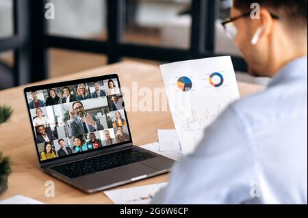 Über die Schulter eines kaukasischen männlichen Büromitarbeiters, hält Finanzdiagramme, hat Online-Videokonferenz mit reifen erfolgreichen ceo und multirassischen Kollegen, mit Laptop, sitzen im Büro Stockfoto