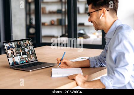 Seitenansicht eines selbstbewussten männlichen Managers, Notizen machen bei Online-Geschäftstreffen mit multinationalen Kollegen, Laptop verwenden, im Büro sitzen, Marketingstrategie planen, Kollegen zuhören Stockfoto