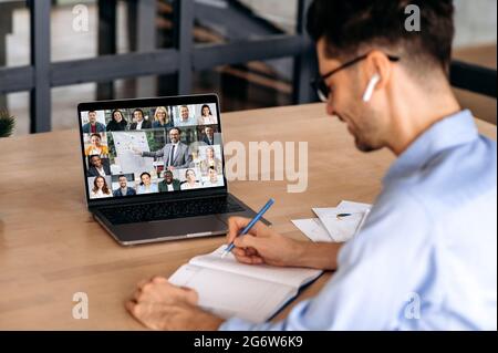 Kaukasischer männlicher Büroangestellter, notiert sich während einer Online-Videokonferenz mit reifen erfolgreichen ceo- und Kollegen aus verschiedenen Rassen, mit Laptop, sitzt im Büro, plant Marketingstrategie Stockfoto