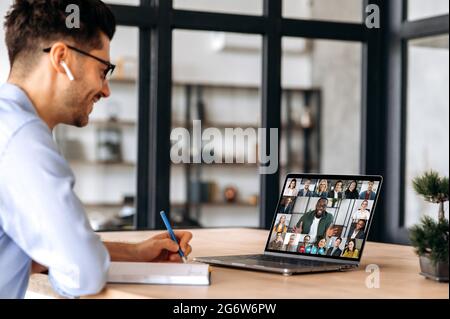 Erfolgreicher männlicher Manager aus Kaukasus, notiert sich während eines Online-Geschäftstreffens mit multinationalen Kollegen, mit Laptop, sitzt im Büro, plant Marketingstrategie, hört Kollegen zu Stockfoto