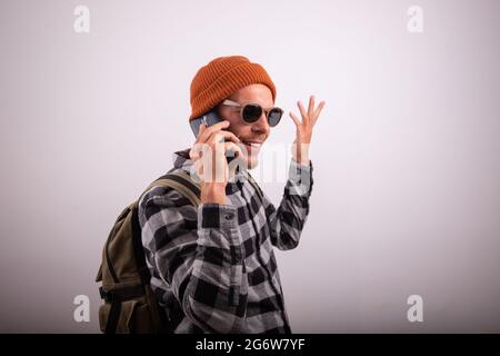 Stilvoller Typ telefoniert, er trägt ein kariertes Hemd, Sonnenbrille und eine Mütze, Hipster mit Rucksack, Foto im Studio mit weißem Hintergrund Stockfoto