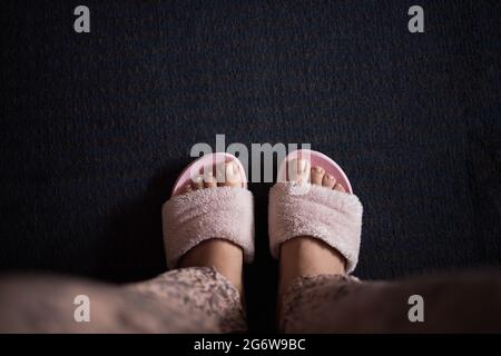 Weibliche Beine mit einer Pediküre in rosa flauschigen Pantoffeln auf einem dunkelblauen Teppich Hintergrund. Kopierraum, flaches Lay, Minimalismus Stockfoto