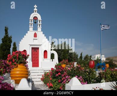 Kleine griechische Kapelle mit Glockenturm und griechischer Flagge umgeben von Blumengarten auf dem Land mit blauem Himmel auf der Insel Mykonos im Frühling, Griechenland Stockfoto