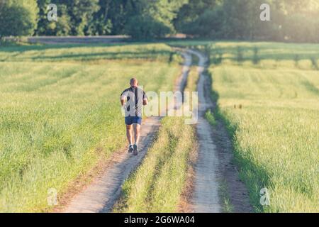 Der sportliche Mann übersteht bei Sonnenuntergang weite Strecken. Der Mensch läuft auf der leeren Landstraße. Stockfoto