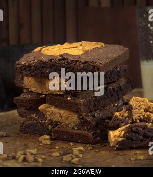 Ein Teller mit frisch zubereiteten Erdnuss- und Keksbrownies auf einem Holzbrett. Mit einem Glas Milch direkt hinter und rechts vom Stapel serviert Stockfoto