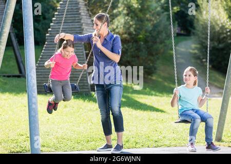 Familie mit zwei Mädchen und Mutter auf Spielplatz Schaukel Spaß haben Stockfoto