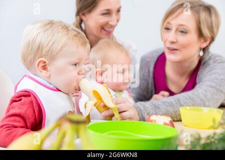 Porträt eines niedlichen und gesunden Mädchens mit blauen Augen, das ihre Mutter anschaut, während sie neben einem anderen Baby zu Hause nahrhaftes Fruchtpüree isst Stockfoto