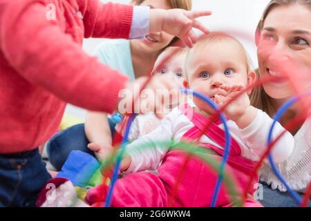 Eine Gruppe von glücklichen jungen Müttern, die ihre süßen und gesunden Babys beobachten, während sie mit bunten Spielsachen in einer modernen Kindertagesstätte spielen Stockfoto