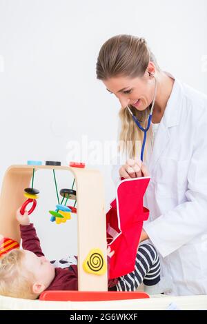 Nettes und gesundes Baby Mädchen spielt mit dem Stethoskop neben ihrer sorgfältigen Mutter, während der routinemäßigen medizinischen Check-up in einer modernen Klinik mit engagierten Stockfoto