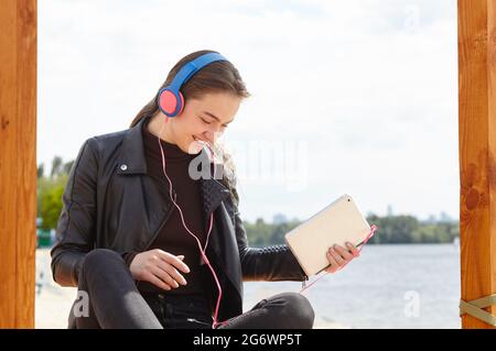 Die junge kaukasische Frau mit blauroten Kopfhörern hört am Flussufer Musik mit einem digitalen Tablet. Portrait Foto von glücklichen Hipster Mädchen gemeinsam Stockfoto