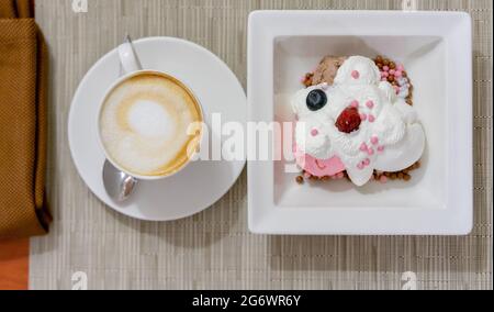 Blick von oben auf Cappuccino in einer weißen Tasse und Vanille- und Erdbeereis mit Schlagsahne und Beeren auf einem weißen Dessertgericht. Stockfoto