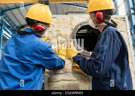 Zwei asiatische Arbeiter, die Isoliermaterial auf einen industriellen Dampfkessel auftragen Stockfoto