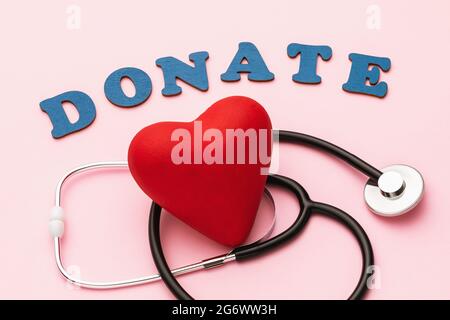 Das Wort aus Holzbuchstaben, einem Spielzeug-Herz und einem medizinischen Stethoskop auf rosa Hintergrund, Konzept zum Thema Spende für die Behandlung Stockfoto
