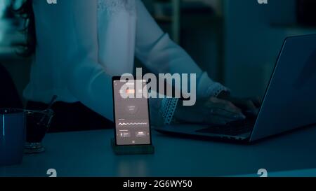 Frau tippt am Laptop, der zu Hause mit einem automatischen Beleuchtungssystem sitzt, das per Sprachsteuerung auf dem Smartphone die Glühbirnen anmacht. Lautsprecher-Gadget reagiert auf Befehle, Person, die das Umgebungslicht steuert Stockfoto