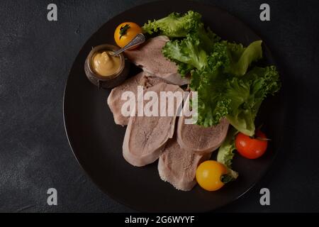 Geschnittene Rinderzungenscheiben auf einer Platte mit Salatblättern, Kirschtomaten und Dijon-Senf auf schwarzem Hintergrund Stockfoto