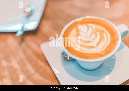 Latte Art in Cappuccino-Kaffeetasse am Kaffeetisch. Nahaufnahme der rosetta-Blumenzeichnung in Schaumstoff. Nahaufnahme einer weißen Kaffeetasse mit herzförmiger Latte-Kunst Stockfoto