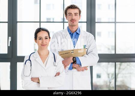 Porträt zweier entschlossener Ärzte, die weiße medizinische Kleider tragen, während sie in einem modernen Gesundheitszentrum mit ernsthaftem Gesichtsausdruck auf die Kamera blicken Stockfoto