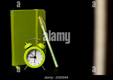 Hellgrüner Wecker mit Stiftnotizbuch auf schwarzem dunklen Hintergrund mit weißem Streifen. Wake-up-Alarm-Konzept. Morgendliche Routine und Produktivität. Zurück zu Stockfoto
