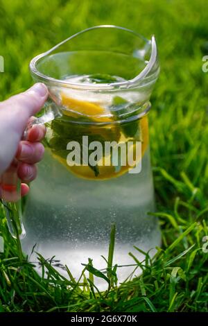 Unschärfe-Hand hält Glaskrug Limonade mit Zitrone und Minze auf natürlichem grünen Hintergrund. Krug mit kalten Sommercocktails oder Punsch im Freien, outsi Stockfoto