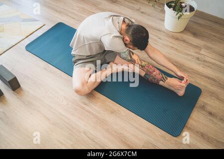 Draufsicht des tätowierten jungen Mannes, der sich auf der Yogamatte dehnt und mit den Händen nach Zehen greift Stockfoto