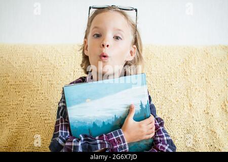 Überraschendes Mädchen in einem karierten Hemd und einer Brille hält einen Stapel Bücher in ihren Händen. Online-Lernkonzept. Stockfoto