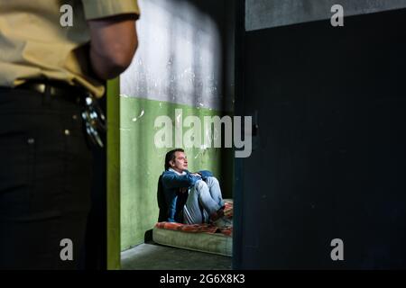 Seitenansicht eines jungen männlichen Gefangenen, der denkt, während er allein auf einer schmutzigen Matratze in einer veralteten Gefängniszelle sitzt, die von einem Polizisten bewacht wird Stockfoto