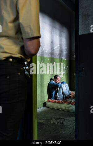 Seitenansicht eines jungen männlichen Gefangenen, der denkt, während er allein auf einer schmutzigen Matratze in einer veralteten Gefängniszelle sitzt, die von einem Polizisten bewacht wird Stockfoto