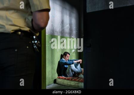 Seitenansicht eines jungen männlichen Gefangenen, der denkt, während er allein auf einer schmutzigen Matratze in einer veralteten Gefängniszelle sitzt, die von einem Polizisten bewacht wird Stockfoto