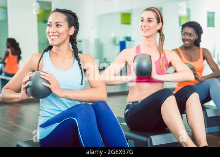 Schöne junge Frau, die während des Gruppenträgertrainings für Frauen in der Turnhalle mit dem med Ball aus der Sitzposition für starke bauchmuskeln den russischen Twist trainiert Stockfoto