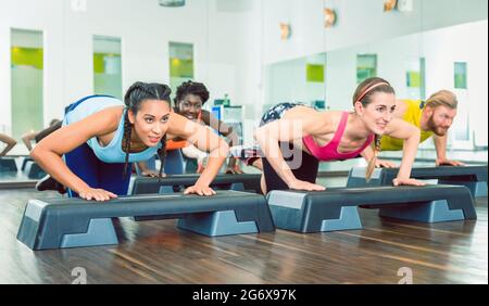 Entschlossene junge Frauen trainieren Liegestütze auf Aerobic-Stepper-Plattformen während des Gruppentraubungstrainings in einem modernen Fitnessclub Stockfoto