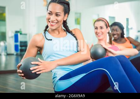 Schöne junge Frau, die während des Gruppenträgertrainings für Frauen in der Turnhalle mit dem med Ball aus der Sitzposition für starke bauchmuskeln den russischen Twist trainiert Stockfoto