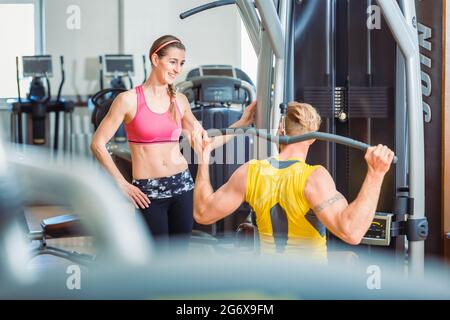 Fit schöne Frau lächelt vor Bewunderung über einen starken Mann Ausübung Overhead lat Pushdown am Kabelgerät in einem trendigen Fitness-Club Stockfoto