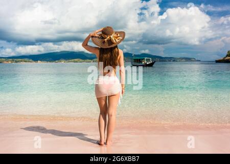 Volle Länge, Rückseite eine attraktive junge Frau mit Strohhut und Mesh schwimmen Rock während Weg am Strand in Insel Komodo, Indonesien suchen Stockfoto