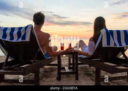 Rückansicht eines verliebten jungen romantischen Paares, das auf Holzstühlen sitzt und während des Urlaubs oder Honigs Cocktails an einem tropischen Strand bei Sonnenuntergang trinkt Stockfoto
