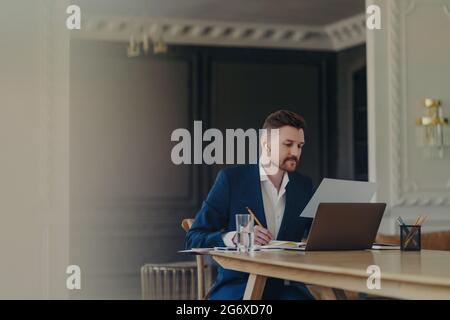 Serious Geschäftsmann in Smart Suit schreibt in Notizblock konzentriert auf Papier Dokument Posen am Schreibtisch mit Laptop-Computer Glas Wasser verwendet Stockfoto