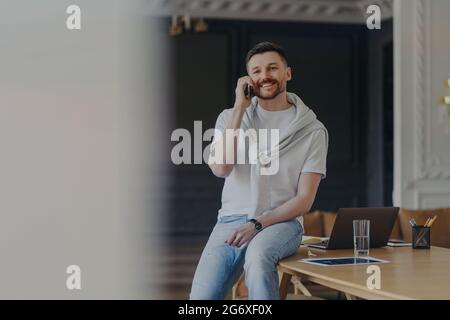 Glücklicher bärtiger männlicher Freiberufler arbeitet im Heimbüro-Raum, gekleidet in legeres T-Shirt und Jeans hat Telefongespräch diskutiert Arbeitsfragen hat Stockfoto