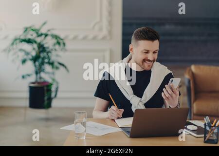 Glücklicher bärtiger junger Mann studiert in Innenräumen mit modernen Gadgets schreibt Notizen von der Internet-Website macht Aufzeichnungen in Notebook hält Smartphone Posen an Stockfoto