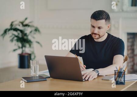 Seriös bärtiger Mann schreibt informative Notizen, während er sich das Webinar auf einem Laptop-Computer ansieht, der in lässigen T-Shirt-Posen am Arbeitsplatz gekleidet arbeitet Stockfoto