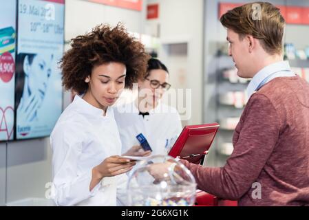 Eine engagierte Apothekerin überprüft ein ärztliches Rezept, während sie verordnete Medikamente an einen jungen männlichen Kunden in einer modernen Drogerie verkauft Stockfoto