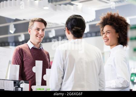Fröhlicher junger Mann und glückliche Kunden im Gespräch mit zwei hilfreiche weibliche Apotheker in einer modernen Apotheke mit freundlichen Angestellten Stockfoto