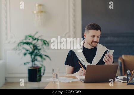 Männlicher Unternehmer, der online studiert und sein Smartphone benutzt, während er an seinem Arbeitsplatz im modernen Büro arbeitet Stockfoto