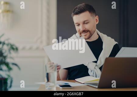 Innenaufnahme des freien männlichen Mitarbeiters konzentriert auf Papiere bereitet Projekt- oder Finanzberichte Posen auf dem Desktop mit Laptop-Computer arbeitet von zu Hause aus Stockfoto